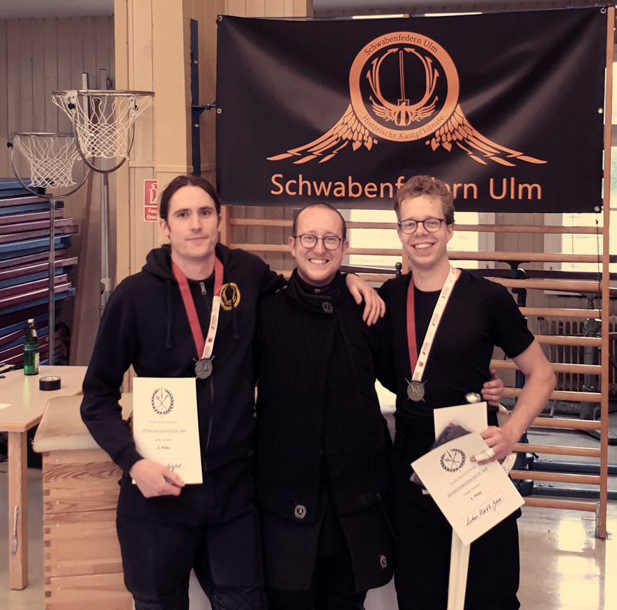 Drei Schwabenfedern nach einem HEMA Schwertkampf Turnier mit ihren gewonnenen Urkunden und Medaillen.
